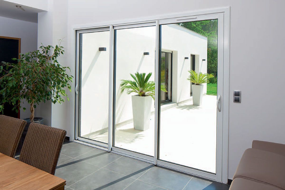 Des portes coulissantes en verre s'ouvrent sur un patio agrémenté de plantes en pot et d'un mur extérieur blanc.
