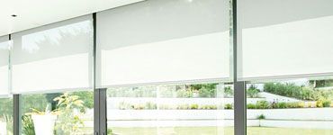 Des stores blancs recouvrent de grandes fenêtres, offrant une vue sur la verdure environnante.