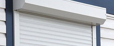 Volet roulant blanc sur une maison, recouvrant partiellement la fenêtre, avec un bardage blanc et des garnitures bleues.