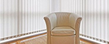 Un fauteuil blanc cassé est placé dans un coin, avec des stores verticaux blancs et un parquet.