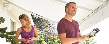 Un couple sous un auvent ; l’homme sourit, tenant un appareil, la femme tient des fruits. En extérieur.