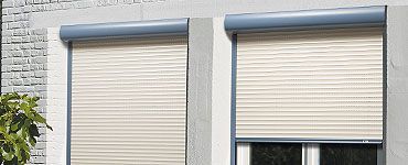 Deux fenêtres avec volets roulants fermés couleur crème ; encadrement bleu.