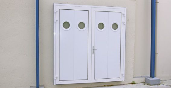 Double porte blanche à fenêtres rondes, encastrée dans un mur beige, avec des tuyaux bleus à proximité.