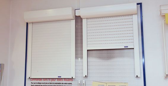 Deux volets roulants blancs, l'un fermé, l'autre partiellement ouvert, fixés au mur.