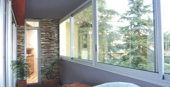 Balcon avec de grandes fenêtres donnant sur les arbres. Mur en pierre, plantes en pot et murs gris.