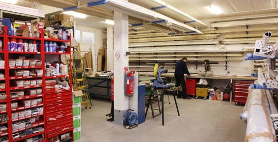 Intérieur d'un atelier avec une personne travaillant à un établi, des étagères à outils et des poutres stockées au-dessus.