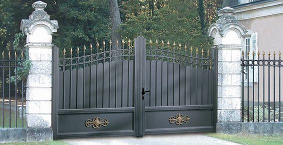 Des grilles en métal noir ouvragé, ornées de détails décoratifs, entre des piliers en pierre et une clôture en fer forgé.