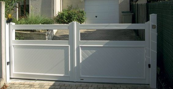 Portail blanc devant une allée, avec une porte de garage en arrière-plan et de la verdure visible.