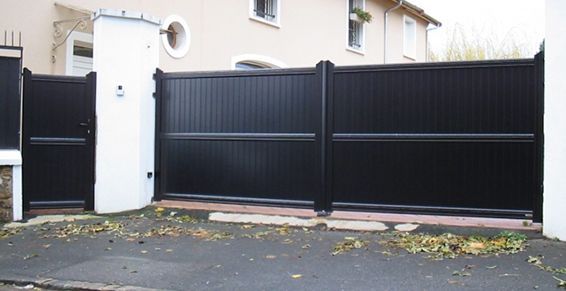 Portail d'allée noir devant une maison, encadré par des piliers blancs.