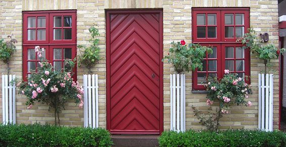 Porte et fenêtres rouges sur un bâtiment en briques beiges, bordé de rosiers et de clôtures blanches.