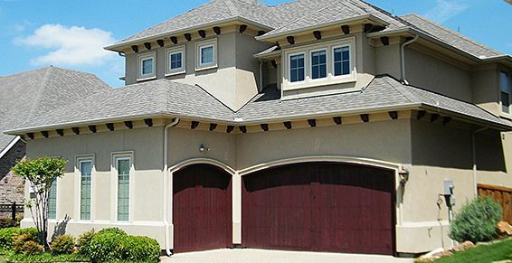 Maison beige à deux étages avec un toit gris et des portes de garage bordeaux, sous un ciel d'un bleu éclatant.