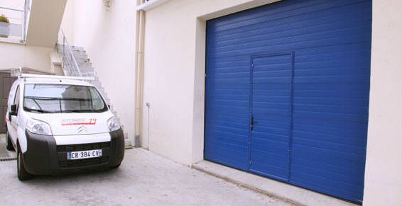 Une camionnette blanche est garée devant une porte de garage bleue. Un escalier se trouve à gauche. L'environnement est lumineux.