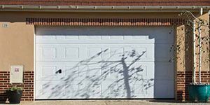 Porte de garage blanche, entourage en briques et l'ombre d'un arbre sur un bâtiment beige.