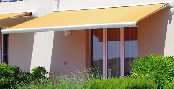 Un auvent jaune s'étend au-dessus d'une fenêtre, adossée à un mur couleur pêche, avec des buissons verts en dessous.