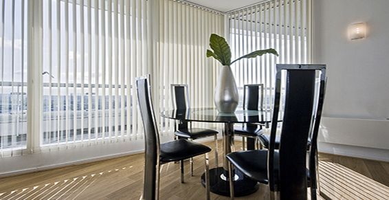 Salle à manger avec table en verre, chaises noires et stores verticaux. Un vase argenté contenant de la verdure est posé sur la table.