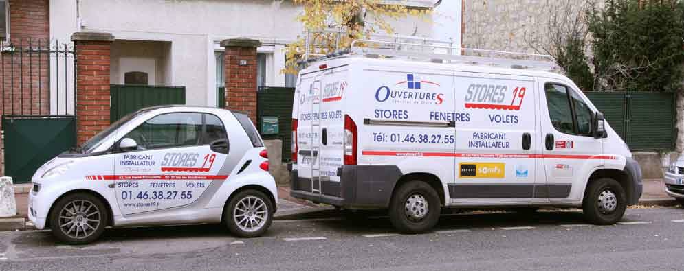 Deux camionnettes blanches arborant des logos d'entreprises sont garées dans une rue près d'un bâtiment au mur de briques rouges.