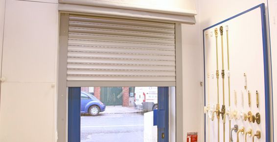 Un rideau métallique masque partiellement une vitrine ; intérieur d'un magasin. Une voiture bleue est visible à l'extérieur.