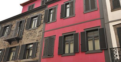Rangée de bâtiments colorés ; façades en pierre, roses et blanches avec des volets sombres.