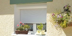 Fenêtre avec volet blanc fermé, paniers de fleurs et fleurs suspendues sur un mur en stuc.