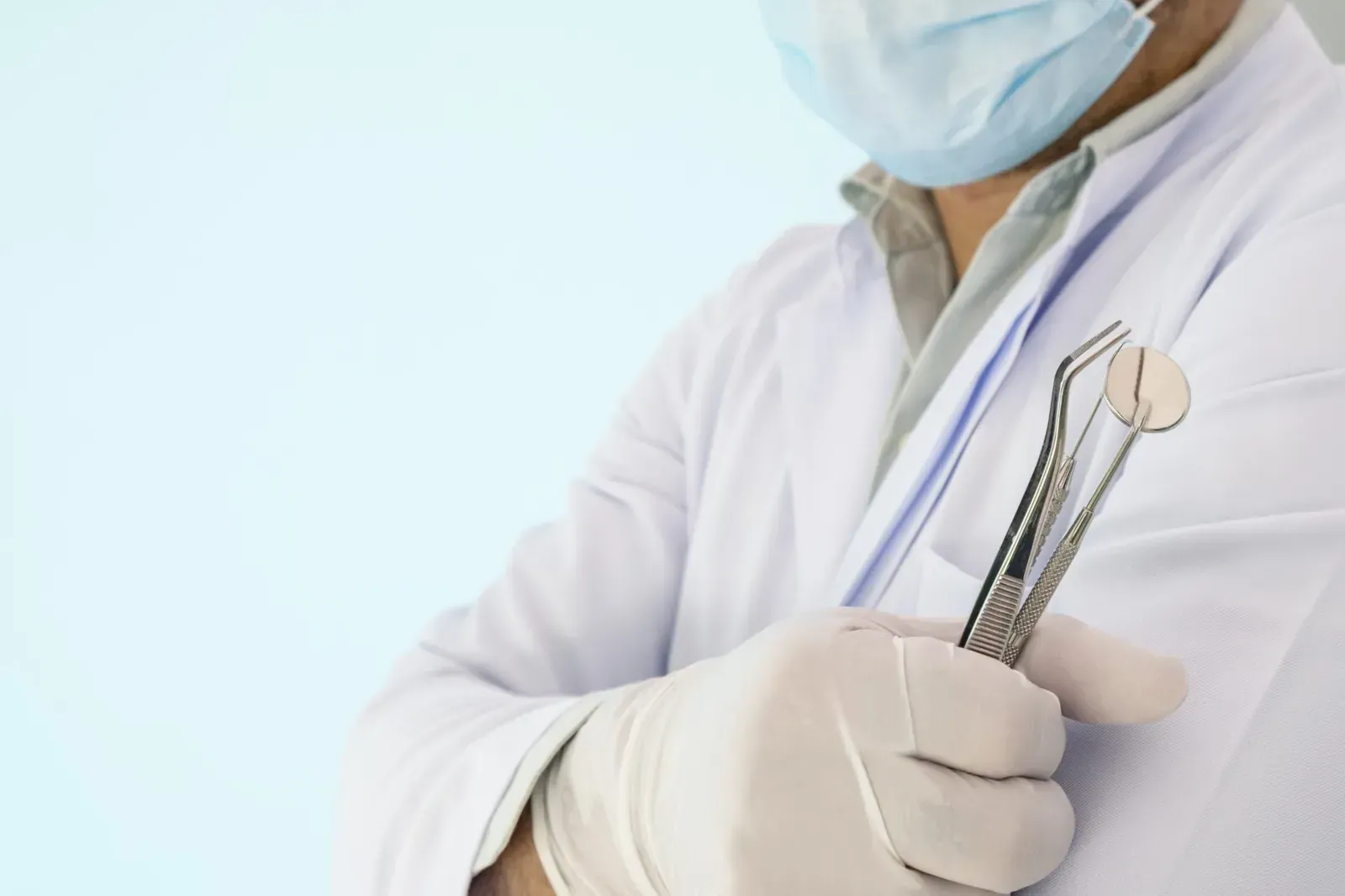 Un dentista con bata blanca, mascarilla y guantes sostiene instrumentos dentales sobre un fondo claro.