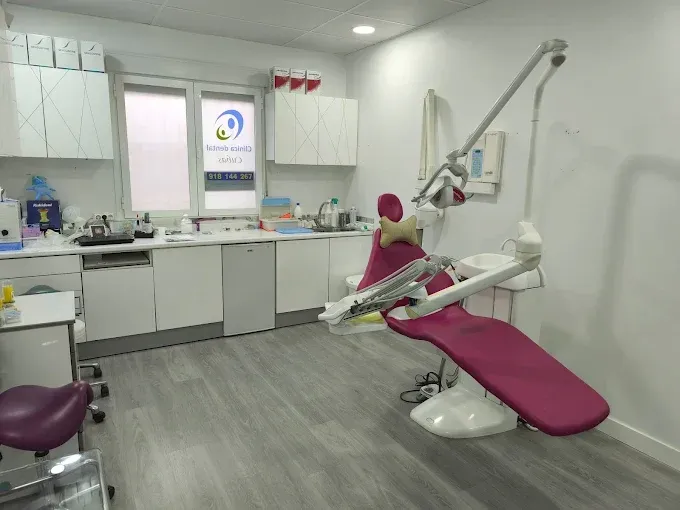 Una sala de exploración dental con un sillón dental rosa, armarios blancos y una ventana con vistas al letrero de la clínica.