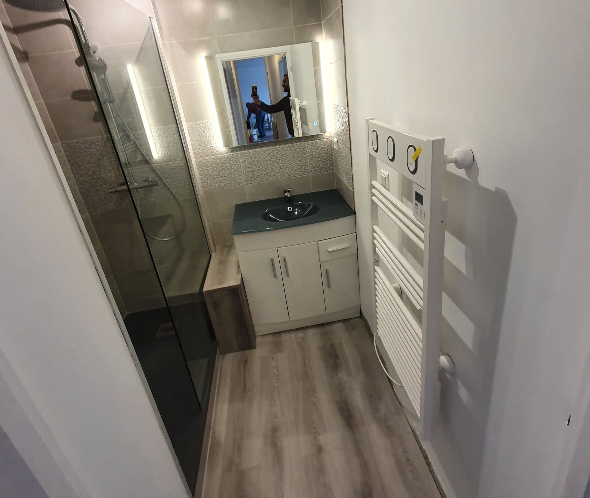 Une petite salle de bains moderne avec une douche vitrée, un meuble-lavabo blanc avec un lavabo foncé et un sèche-serviettes mural.