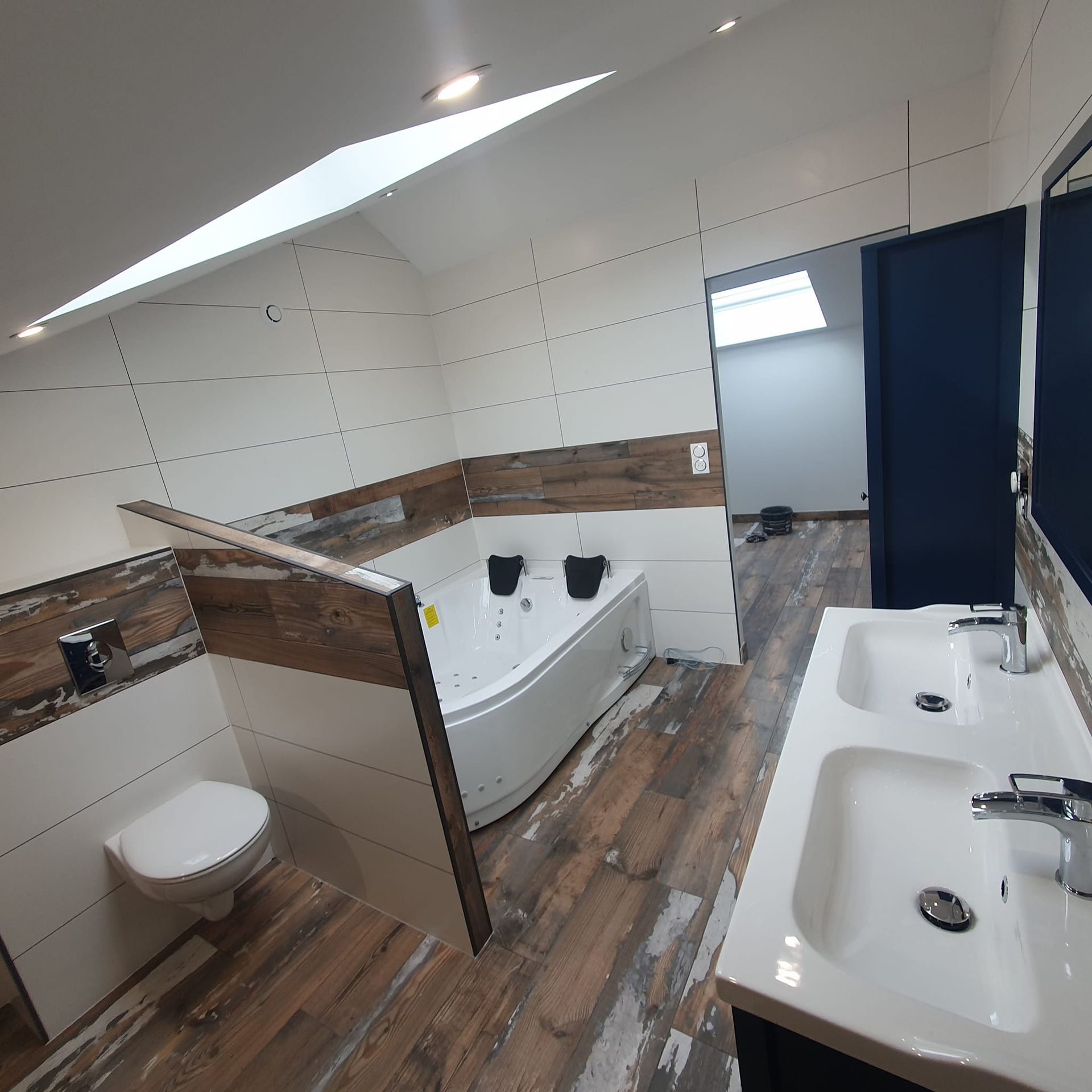 Une salle de bains moderne avec une baignoire d'angle blanche à jets, une double vasque, des toilettes et un sol carrelé imitation bois.