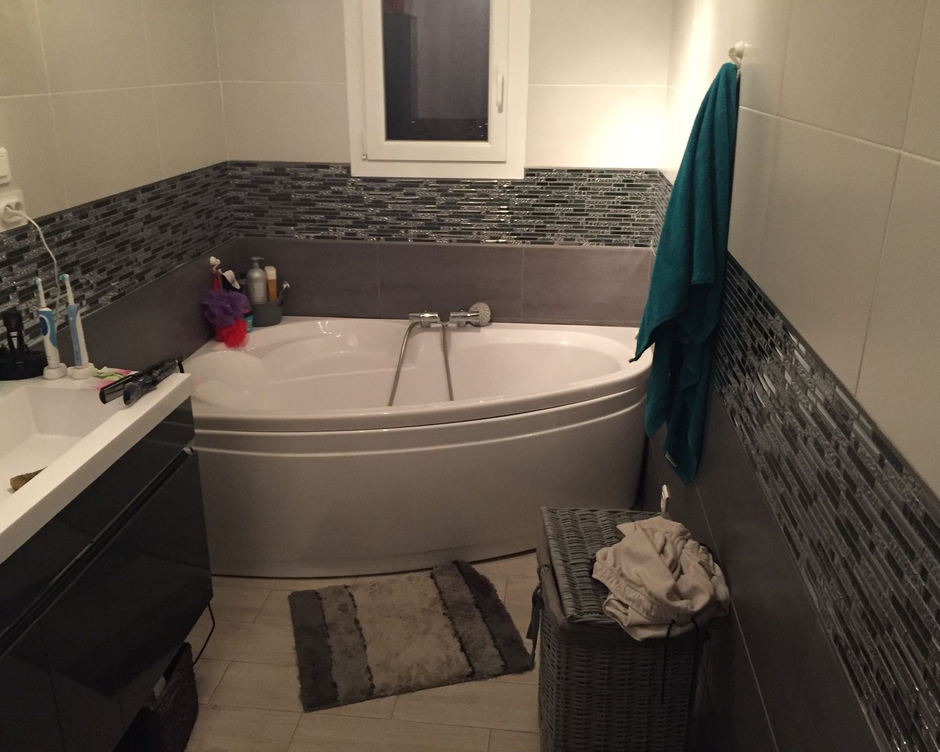 Une salle de bains moderne comprenant une baignoire d'angle blanche, des murs carrelés gris avec une bordure en mosaïque, un meuble lavabo.