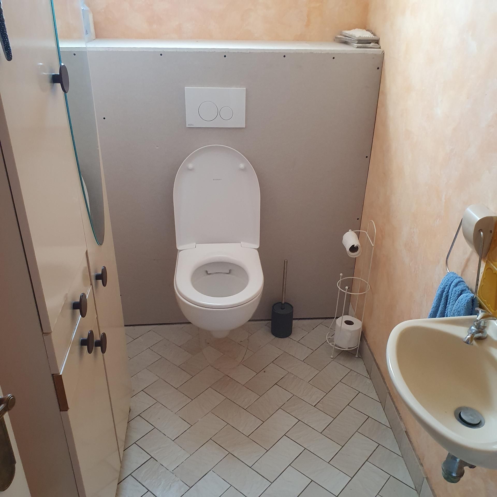 Une petite salle de bains avec des toilettes blanches suspendues, une cloison grise, un lavabo à droite et un sol carrelé en chevrons.