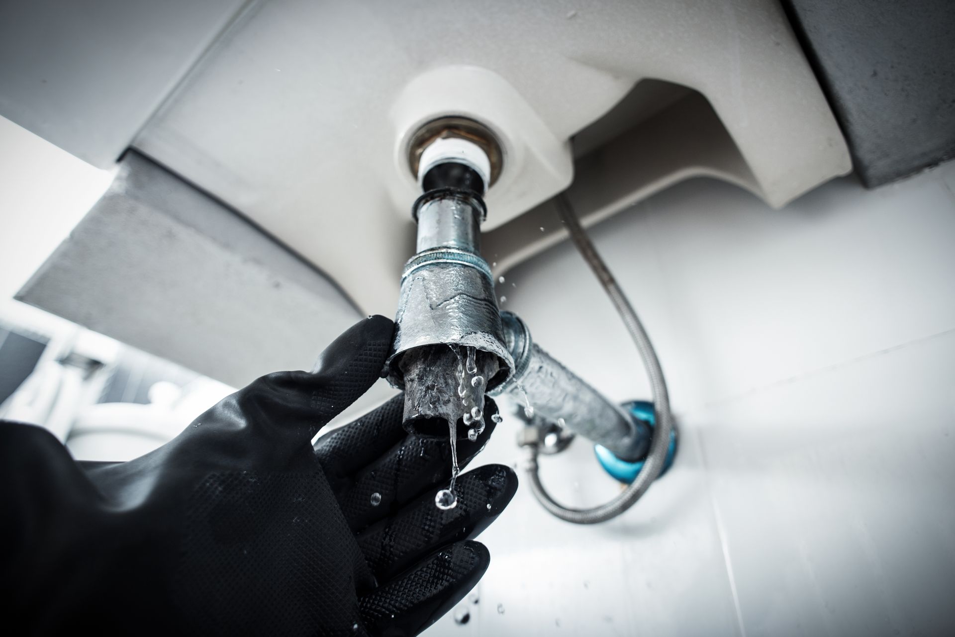 Une personne portant un gant en caoutchouc noir tient un siphon de tuyau d'évier métallique qui fuit sous un lavabo blanc.