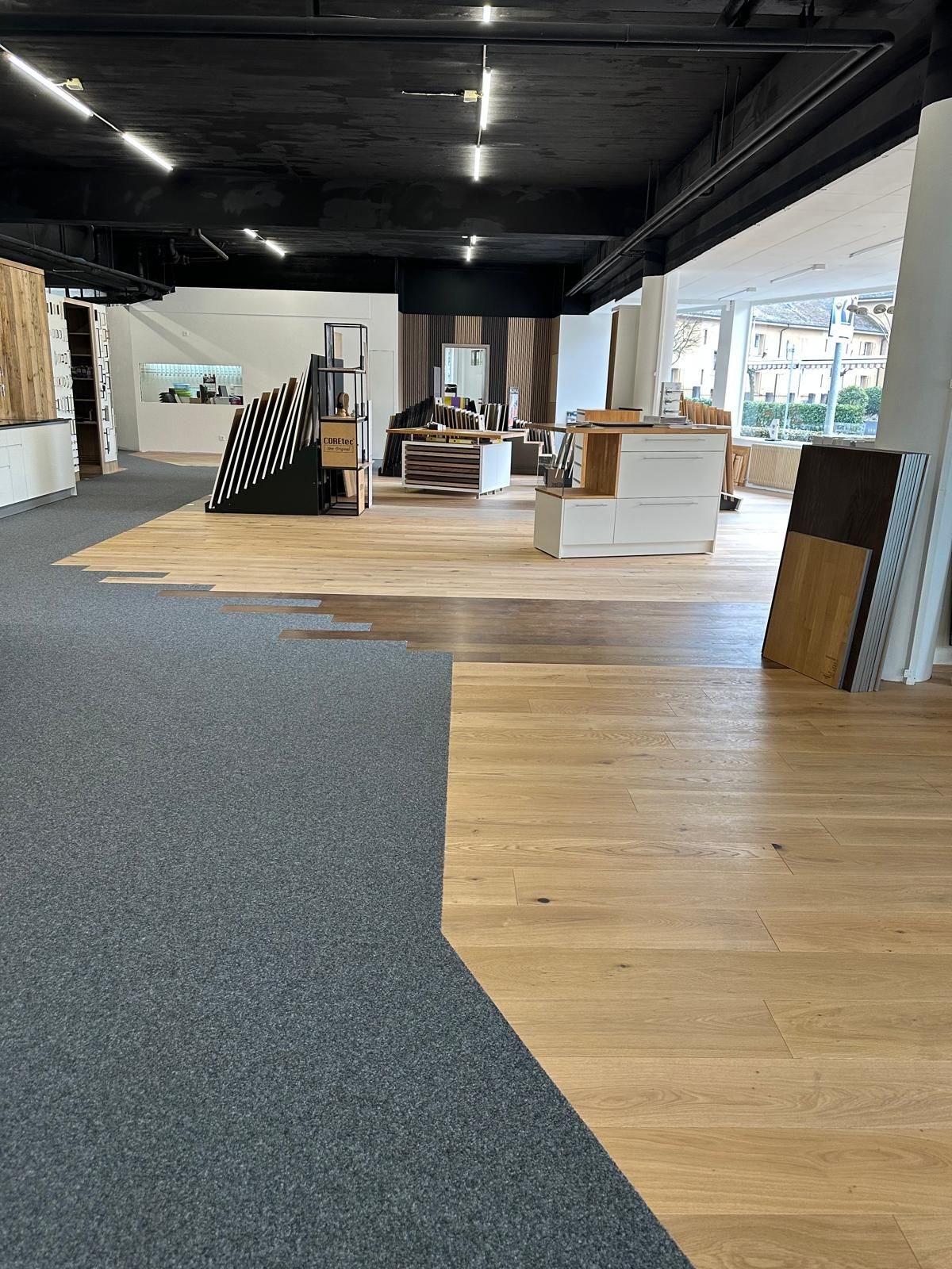 Salle d'exposition spacieuse avec sols en bois et en moquette, meubles exposés et poutres apparentes noires au plafond.