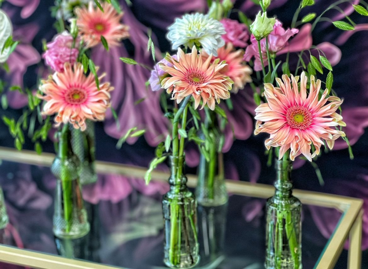 Rosa Gerbera und andere Blumen in Glasflaschen auf einem Tisch vor einem Hintergrund mit Blumenmuster.
