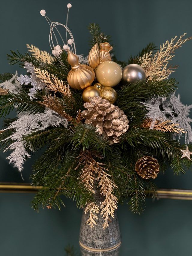 Ein festliches Weihnachtsarrangement in einer Vase. Es besteht aus Tannenzweigen, Gold- und Silberornamenten, Tannenzapfen und dekorativen Zweigen.
