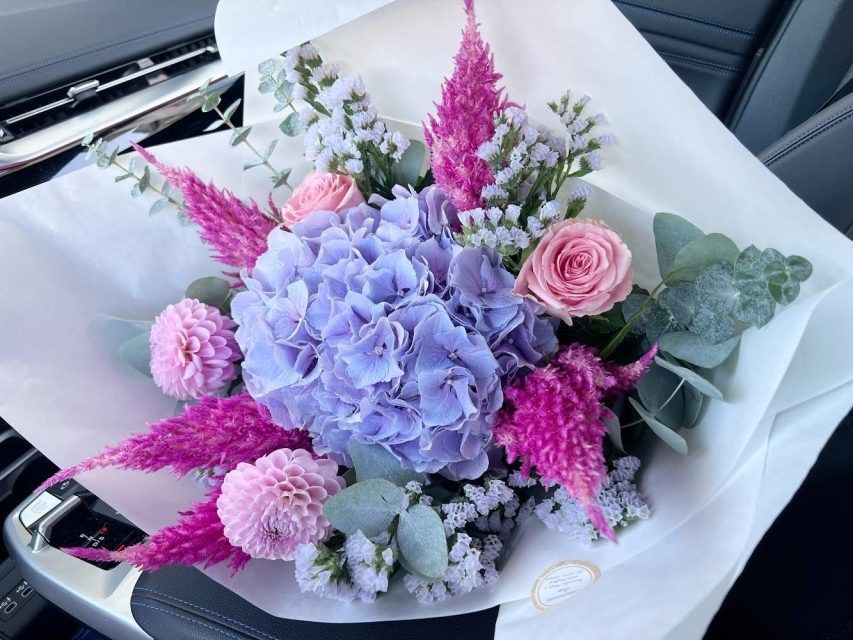 Strauß aus lila Hortensien, rosa Rosen und anderen bunten Blumen, in weißes Papier eingewickelt, in einem Auto.