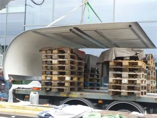 Un camion à plateau transportant des palettes en bois empilées, et une structure blanche incurvée est soulevée au-dessus.