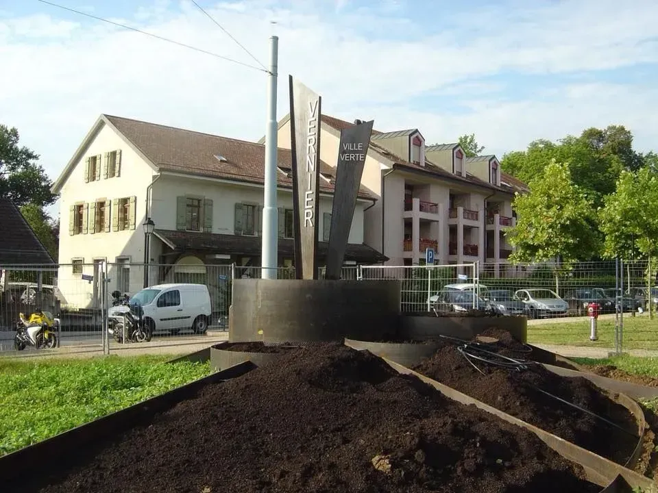 Sculpture dans un espace paysager avec des bâtiments et des véhicules en arrière-plan.