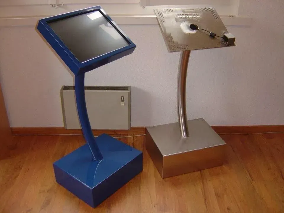 Deux kiosques d'information, l'un bleu avec un écran, l'autre argenté avec une plaque métallique, sur un plancher en bois.