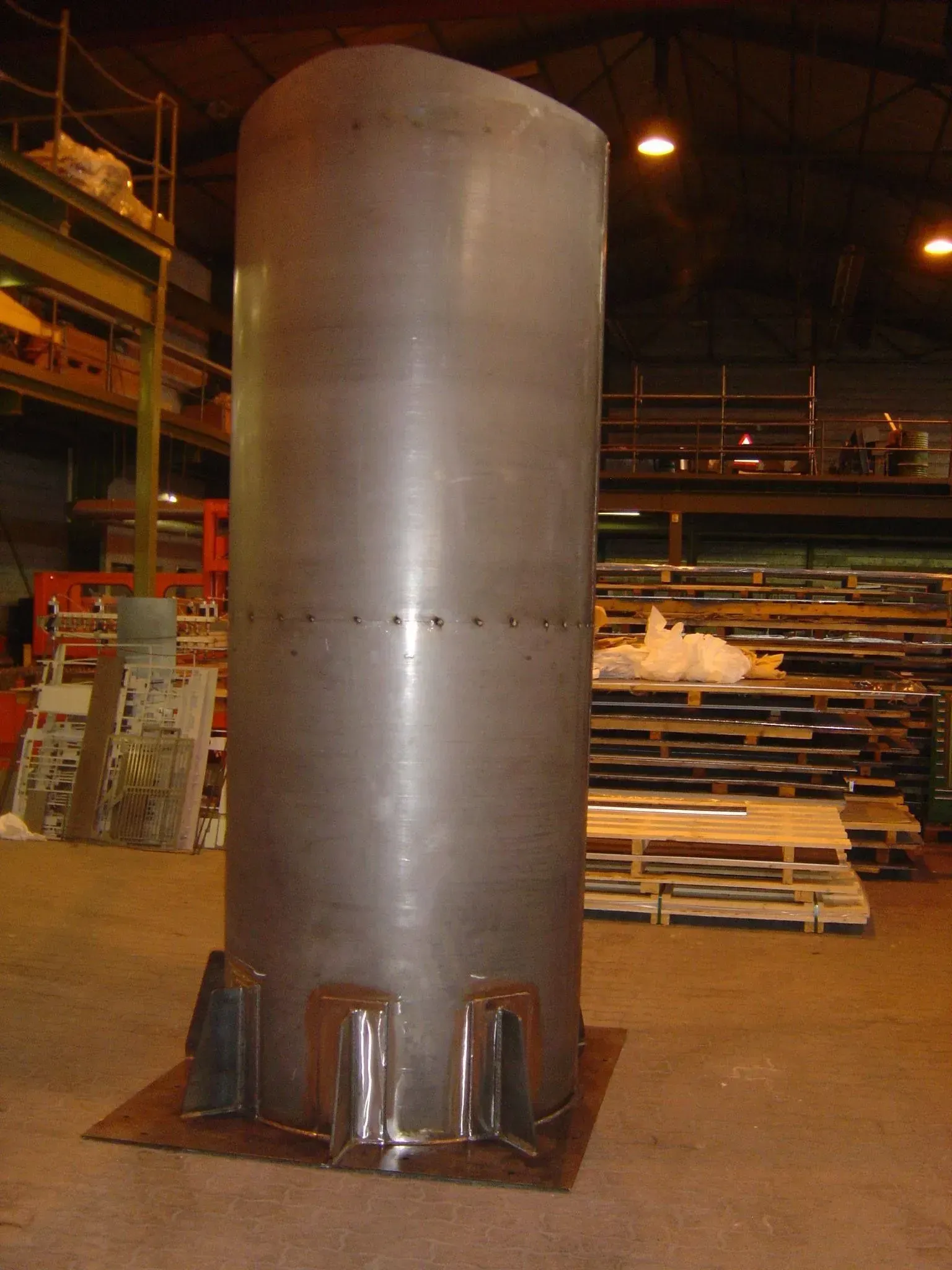 Un grand réservoir métallique cylindrique dans un décor d'usine, posé sur une base métallique avec des pieds de support.