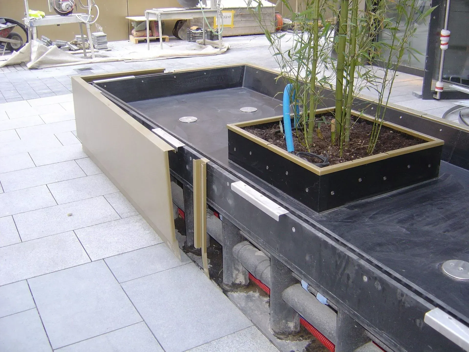 Longue jardinière d'extérieur en bambou, sur une terrasse carrelée. Beige et noir, avec quelques tuyaux visibles.