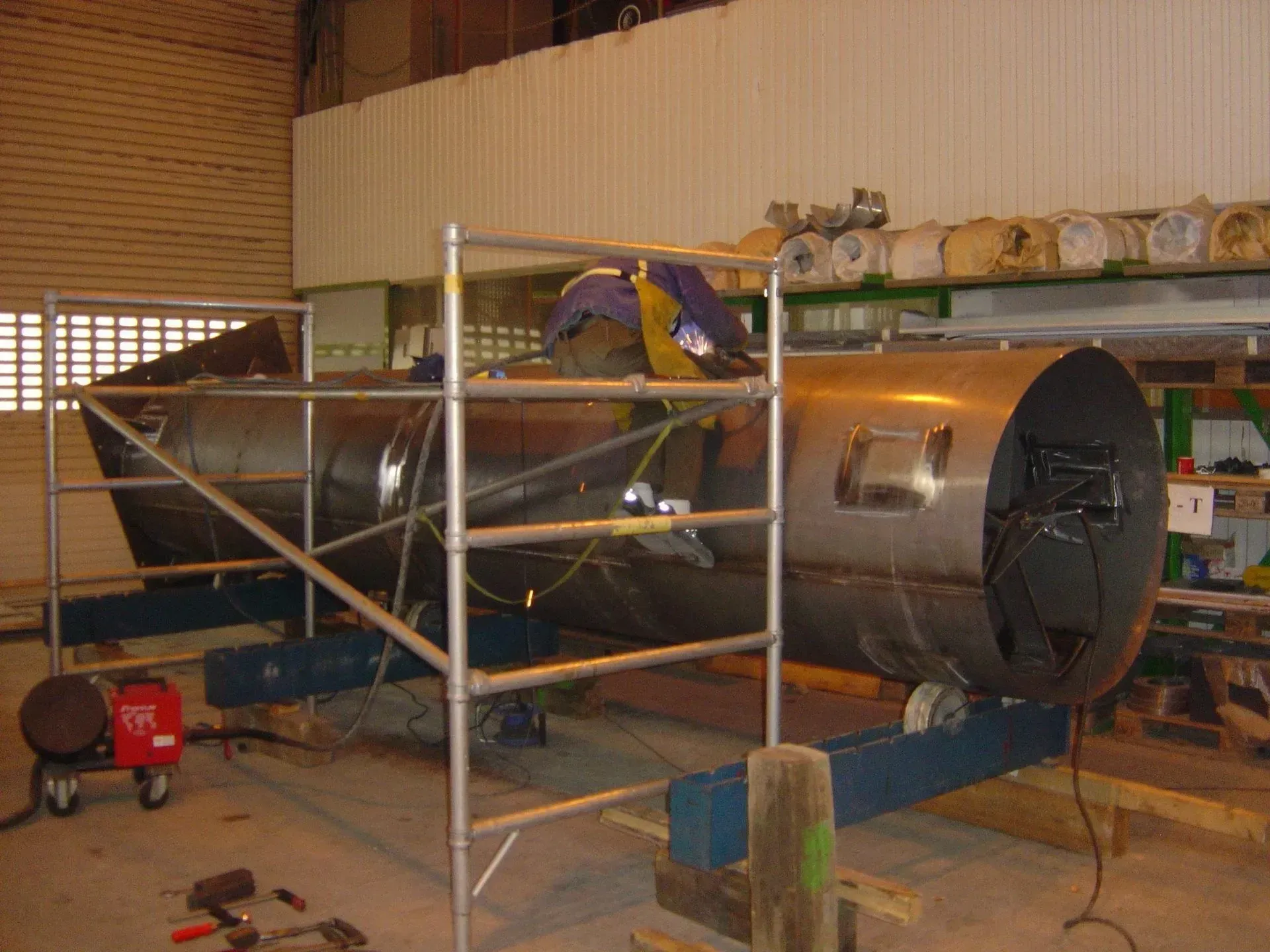 Personne soudant un réservoir métallique, à l'aide d'un échafaudage, dans un atelier.