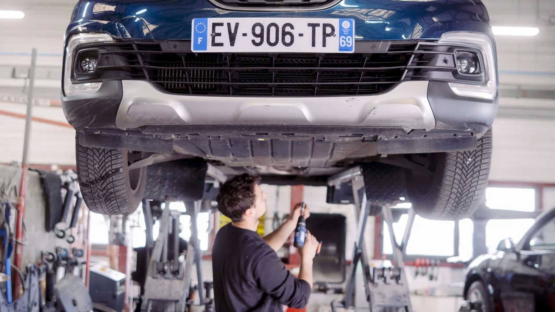 Une voiture bleue est soulevée sur un pont élévateur dans un garage, un mécanicien travaille dessous.