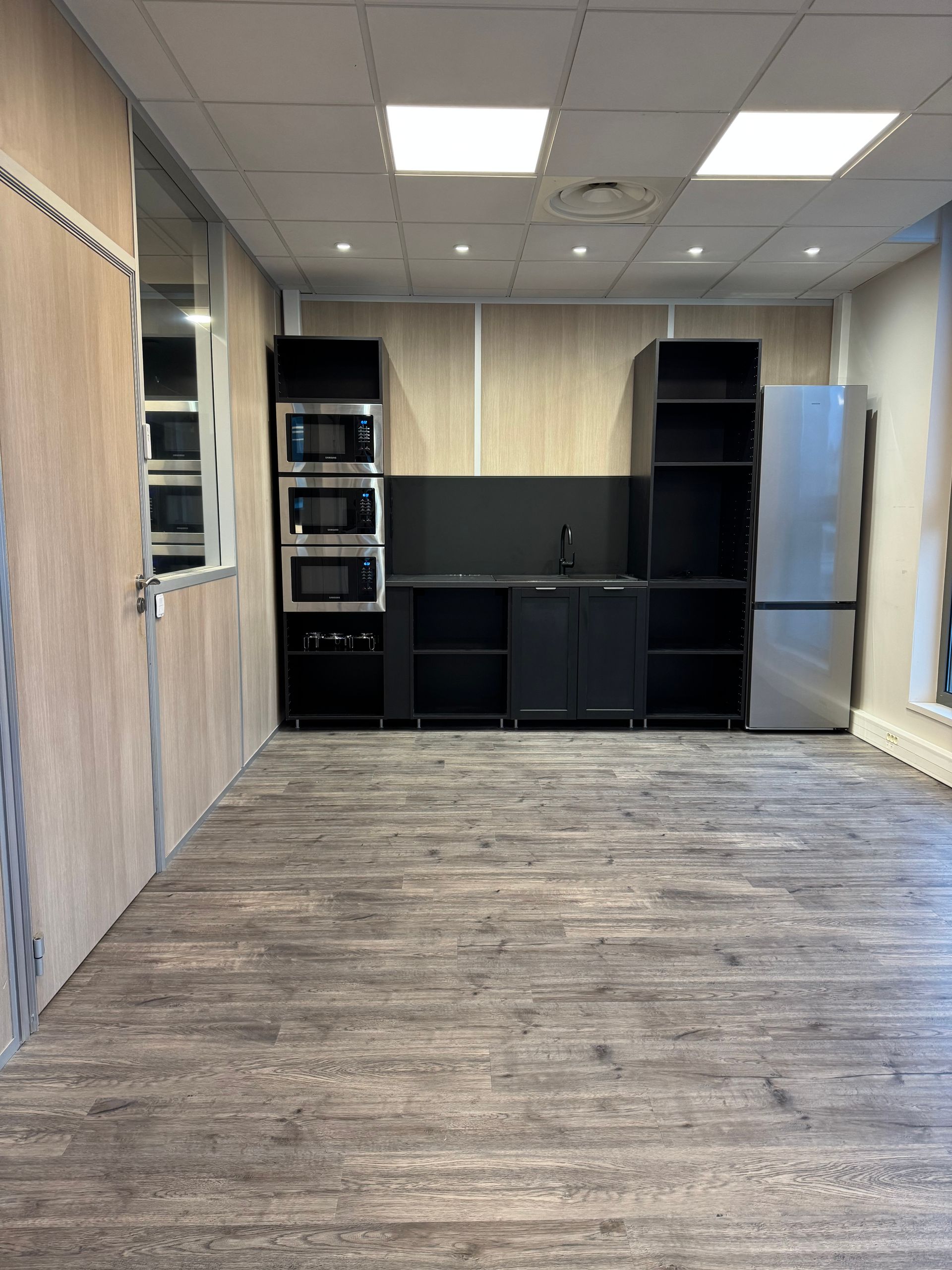 Coin cuisine avec placards noirs, micro-ondes, réfrigérateur et parquet clair.