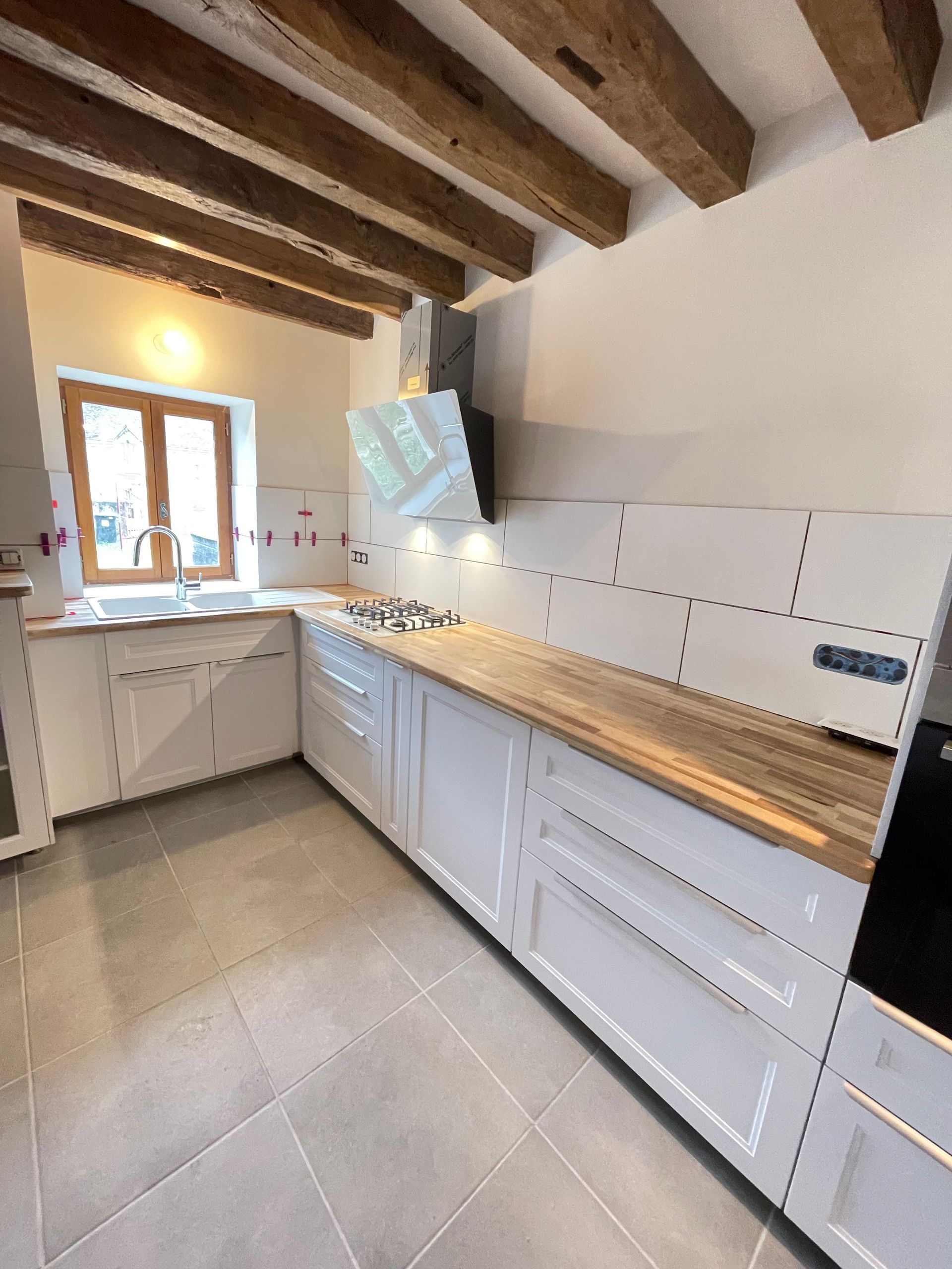 Cuisine blanche avec comptoirs en bois, sol carrelé, poutres apparentes au plafond et une fenêtre.