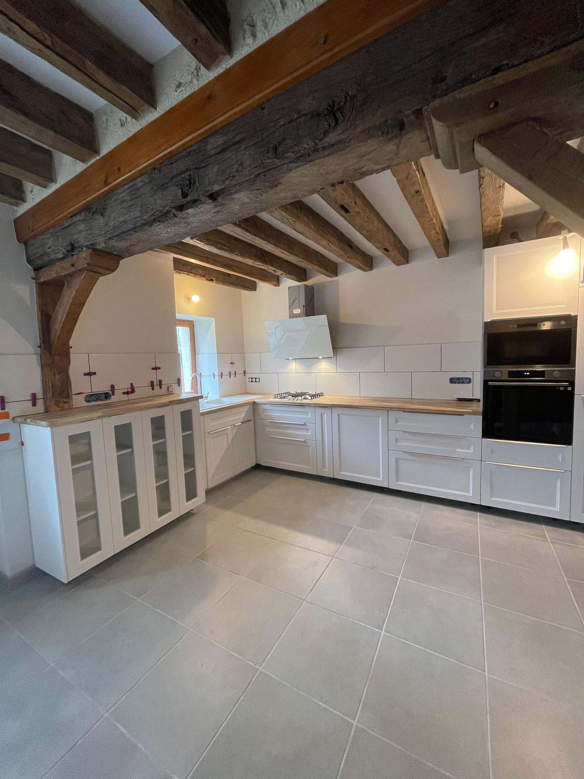 Cuisine avec placards blancs, poutres apparentes, plans de travail en bois clair et sol carrelé gris.