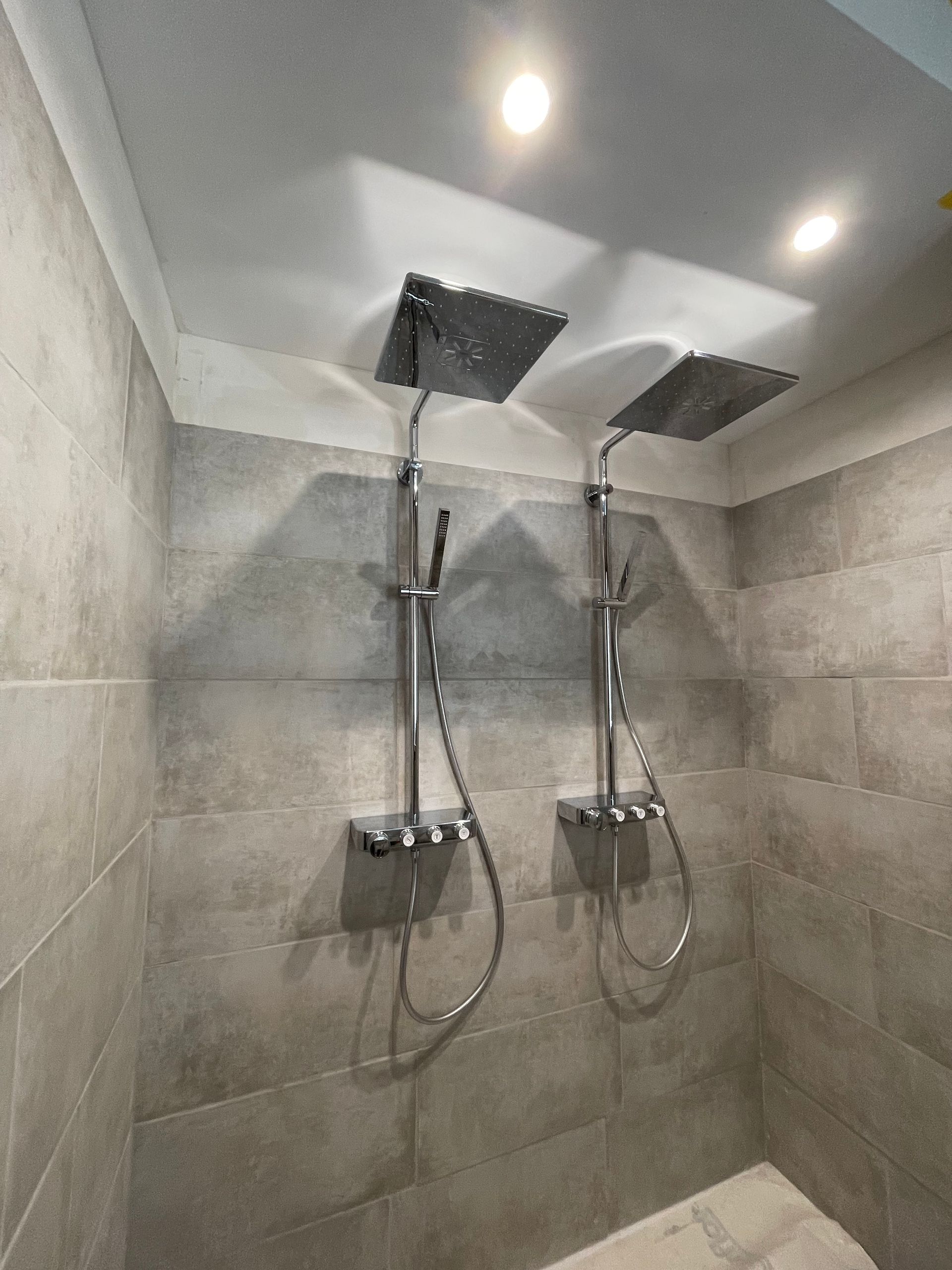 Deux pommeaux de douche argentés avec douchettes, fixés sur un mur carrelé, sous des spots encastrés au plafond.