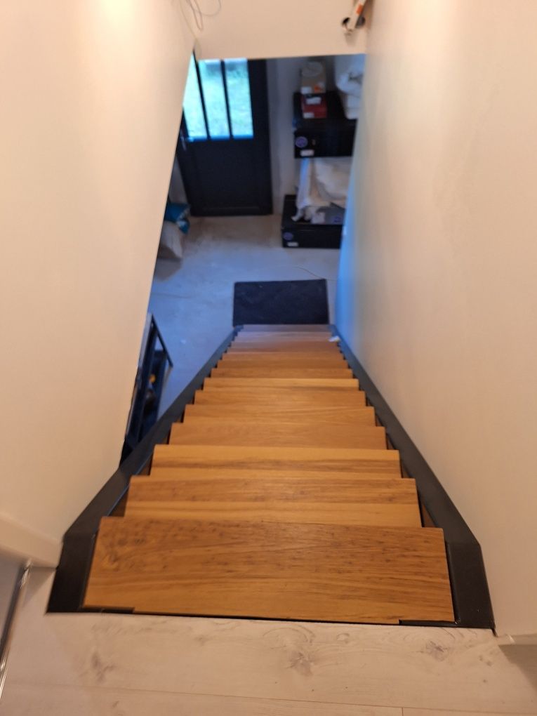 Un escalier en bois descend vers une porte et un espace de rangement. Les murs sont blancs et les marches sont bordées de noir.