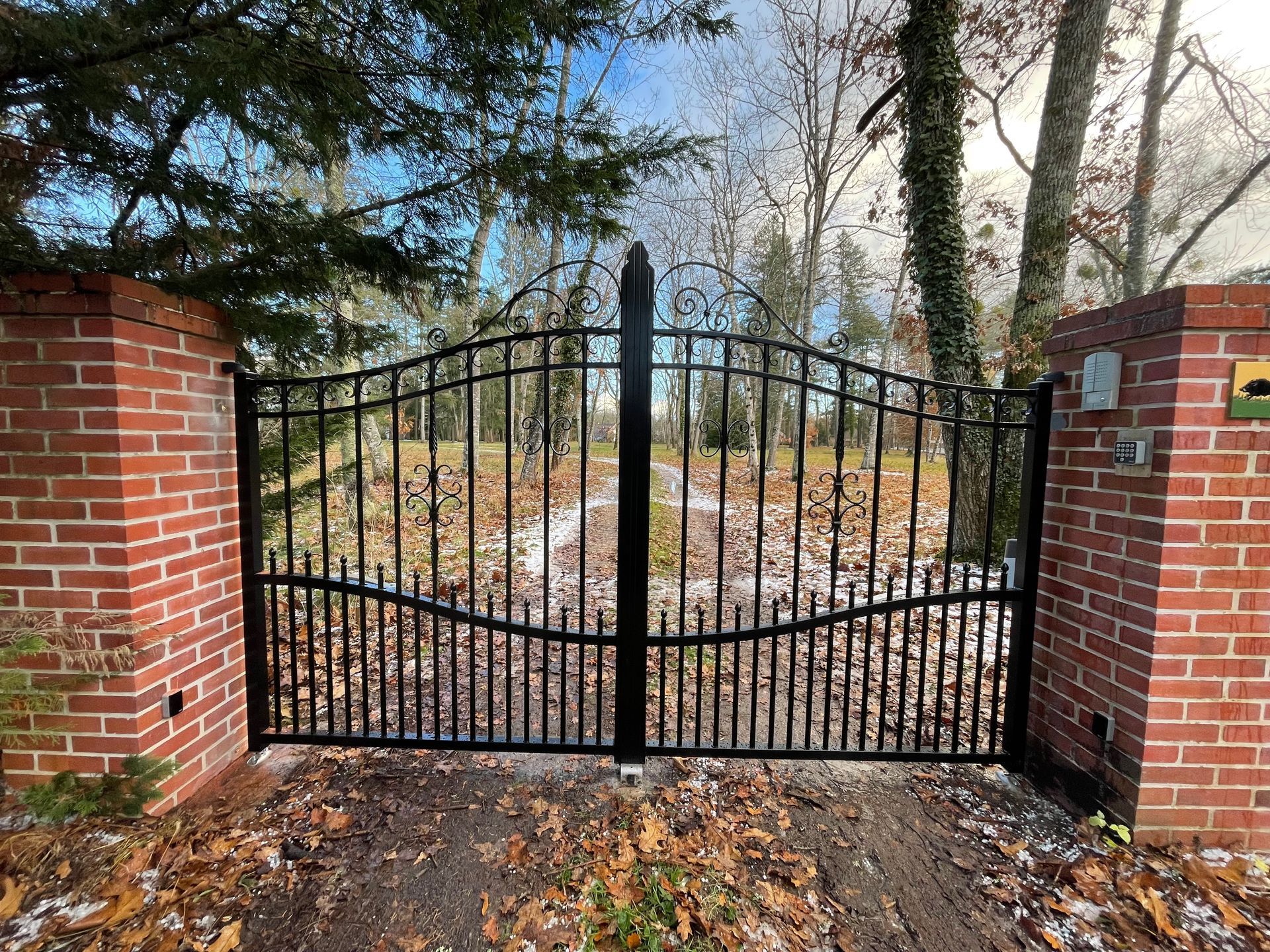 Un portail en fer forgé noir, entre des colonnes de briques, partiellement ouvert sur une zone boisée, avec des feuilles mortes au sol.