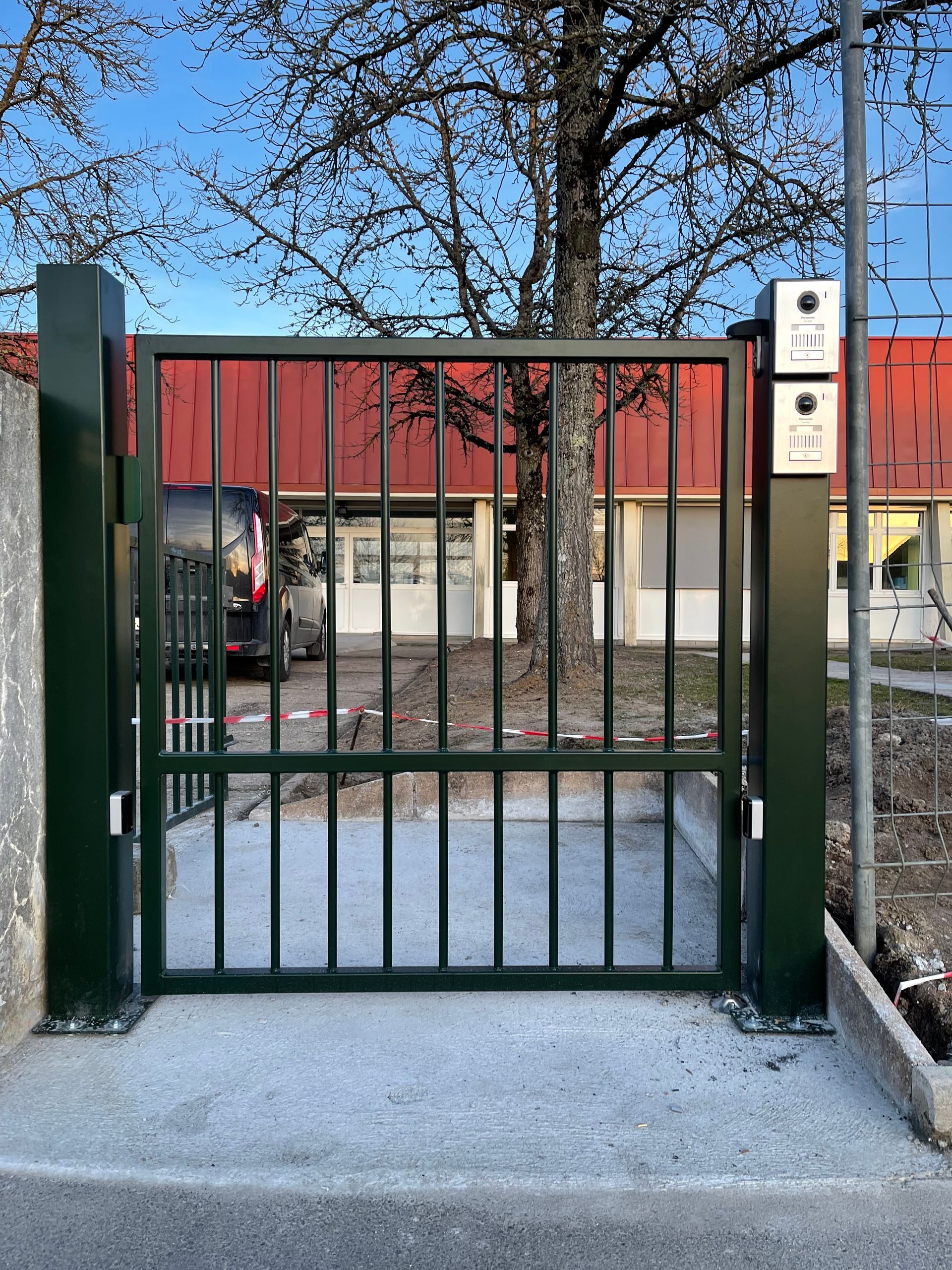 Portail métallique vert à barreaux verticaux donnant sur un bâtiment au toit rouge.