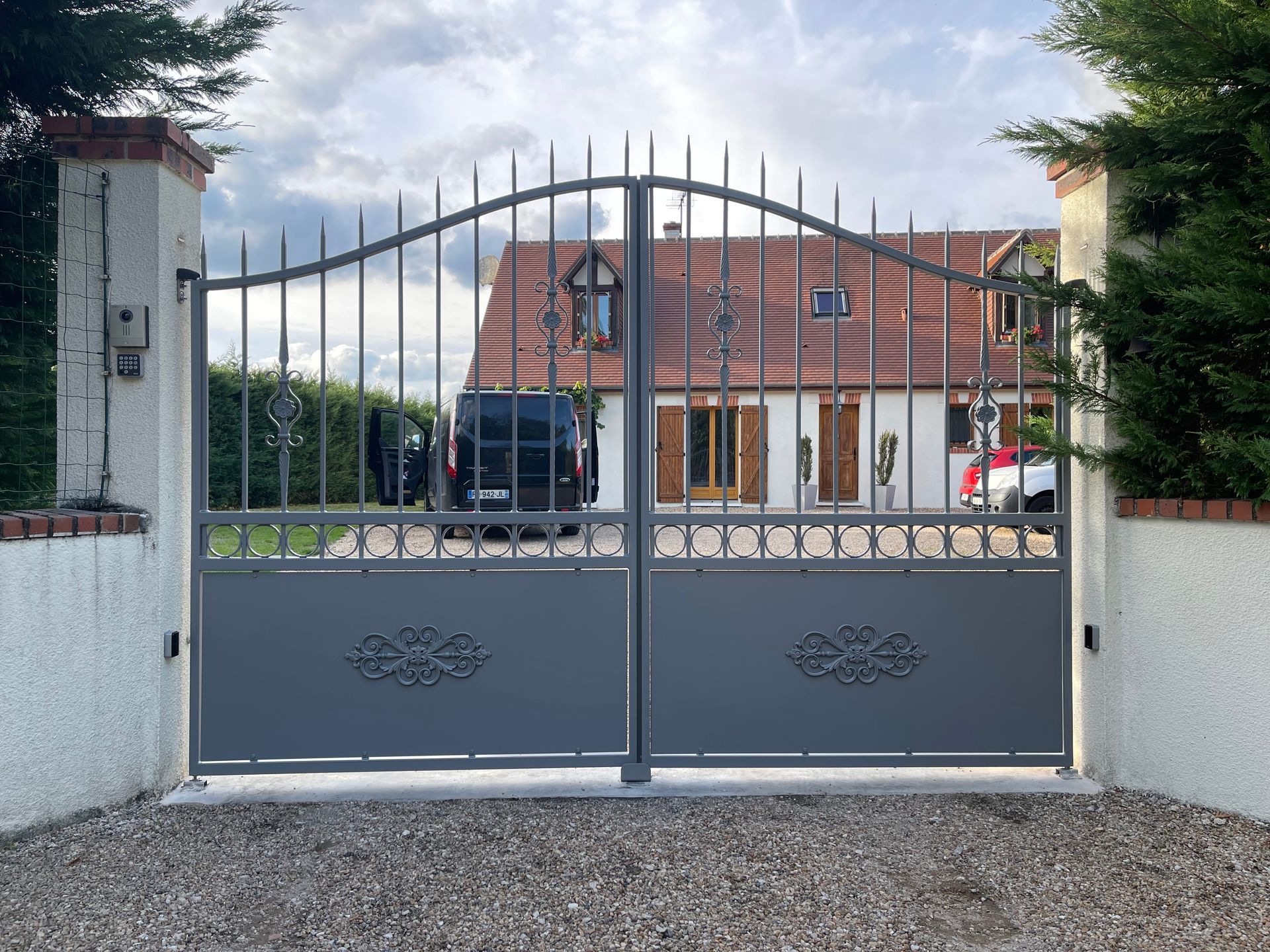 Portail métallique gris devant une maison au toit rouge ; allée, ciel couvert.