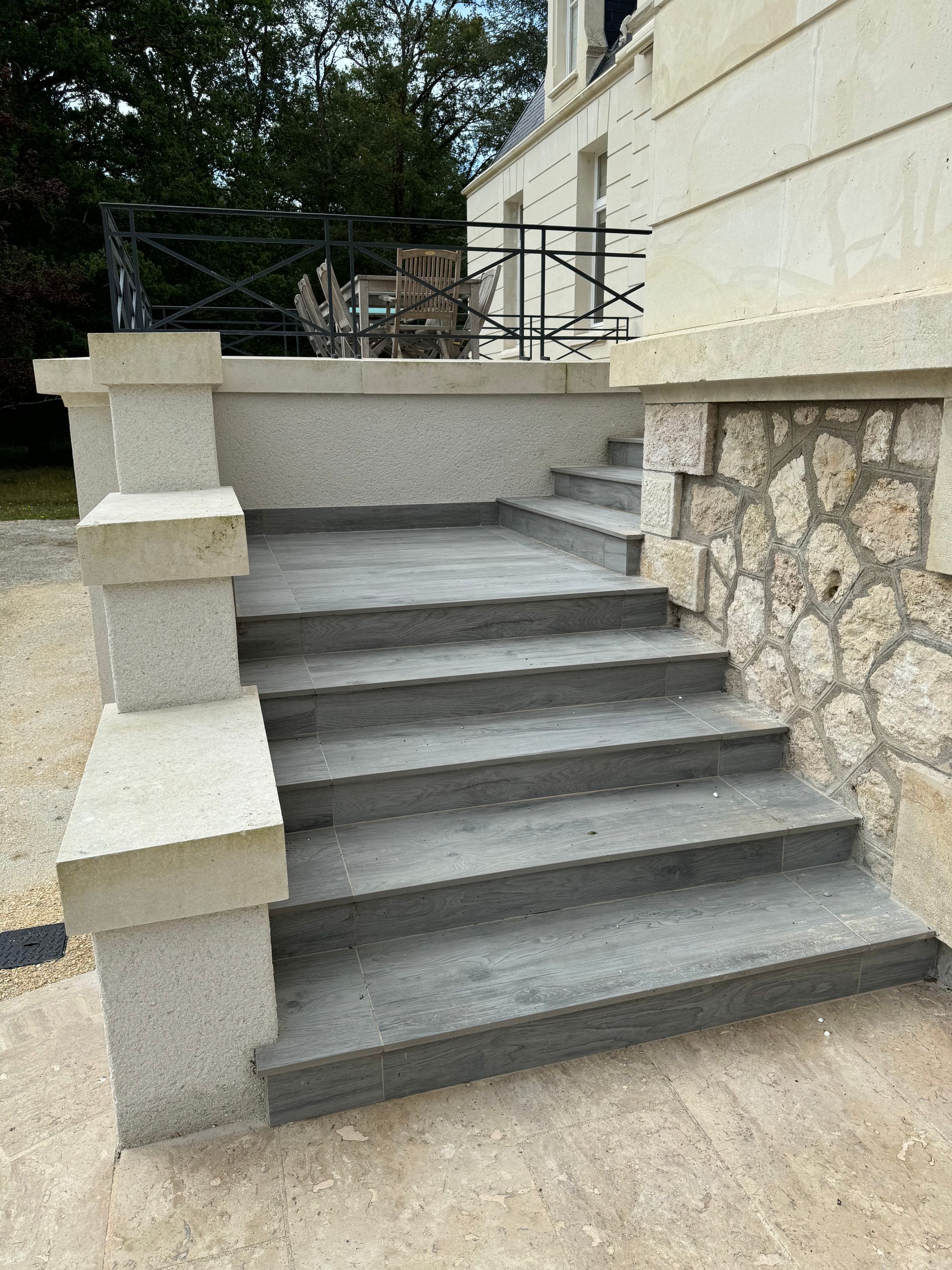 Escaliers en béton menant à l'entrée d'un bâtiment. Mur en pierre à droite, poteaux décoratifs à gauche.